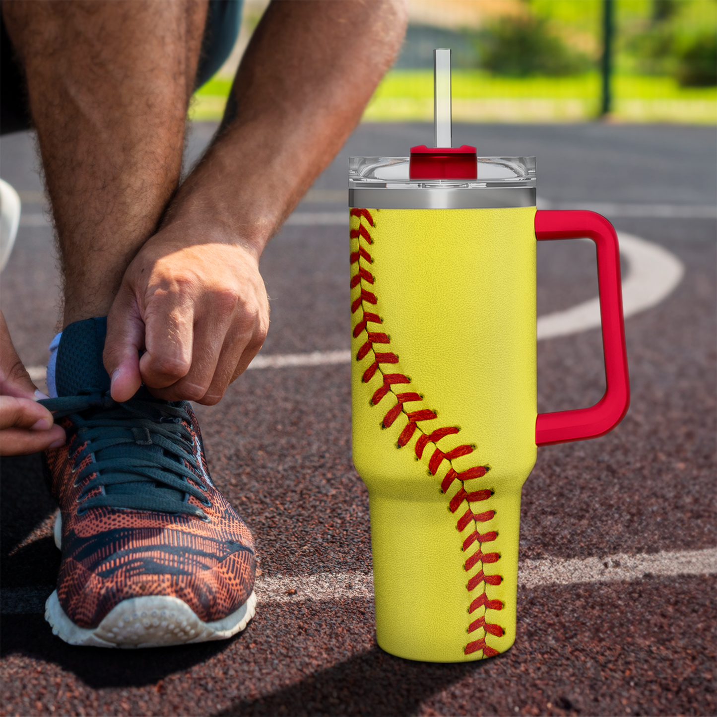 Yellow softball tumbler with red handle and red baseball seam design on outdoor pavement