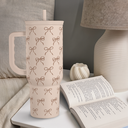 Light tan tumbler with brown bow print, handle and straw, beside a book