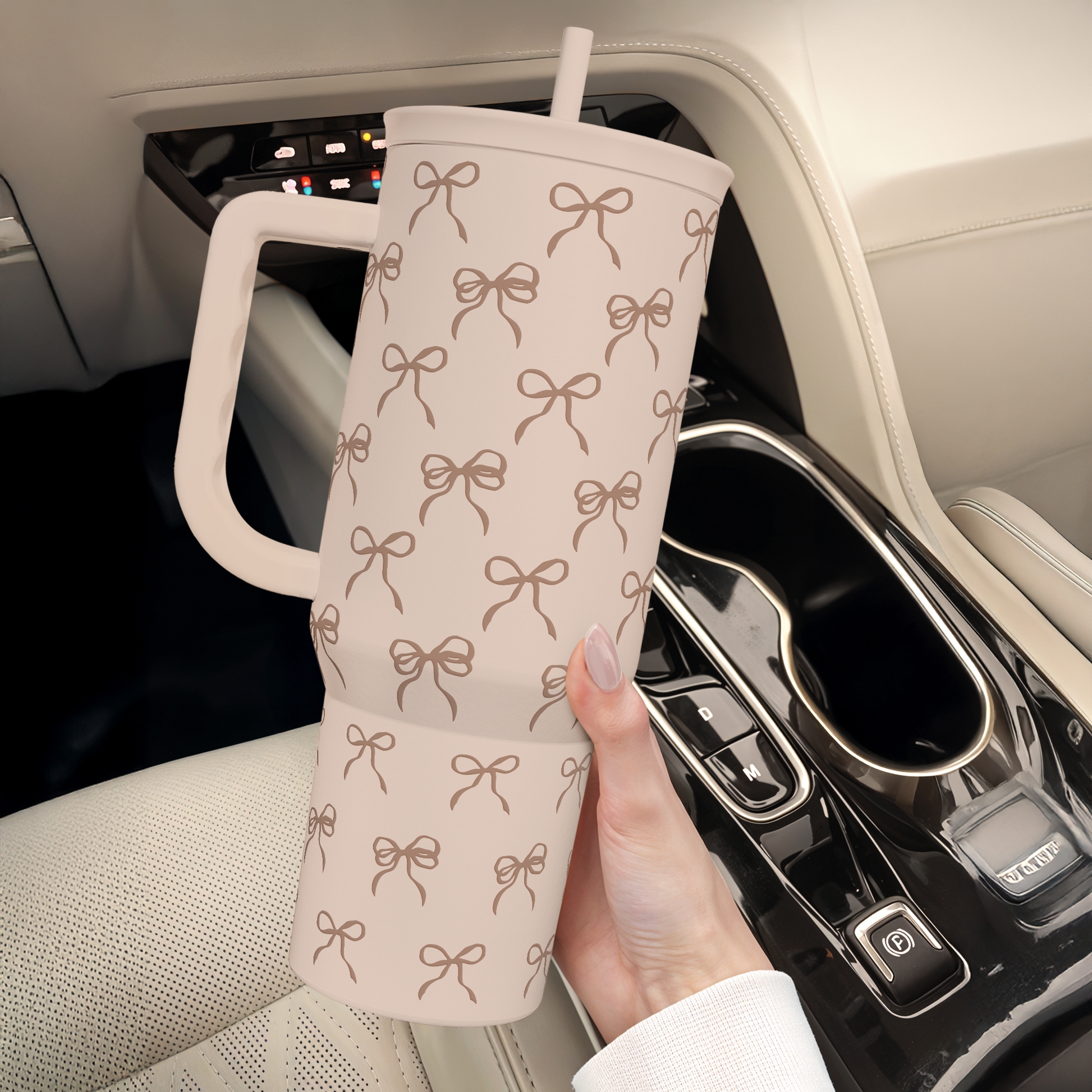 Light tan bow-pattern travel tumbler with handle and straw, held near a car console