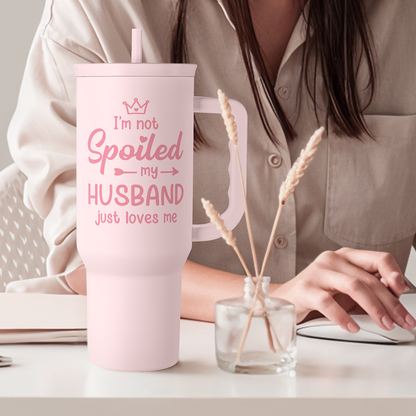 Pink tumbler with straw and handle; “I’m not Spoiled my HUSBAND” slogan on front