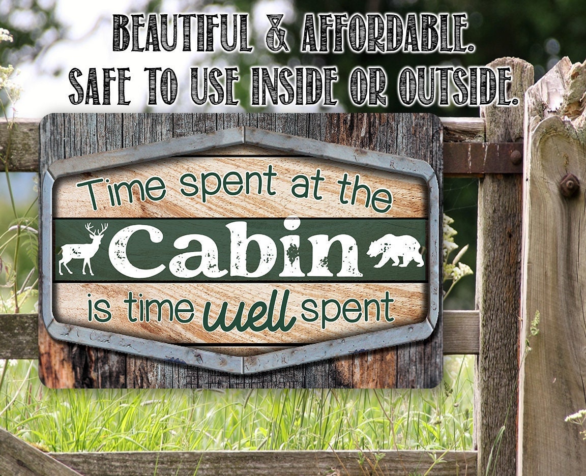 Cabin metal sign outdoors with green panel, “Cabin” lettering, deer silhouettes, and border